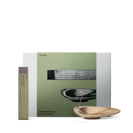 An incense stick rests in a bronze-coloured holder beside a tall packet, in front of an olive-green Aesop gift box.