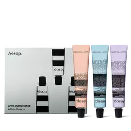 Three Aesop hand balm tubes in peach, pale blue and lilac stand on black caps beside an illustrated silver-grey box.