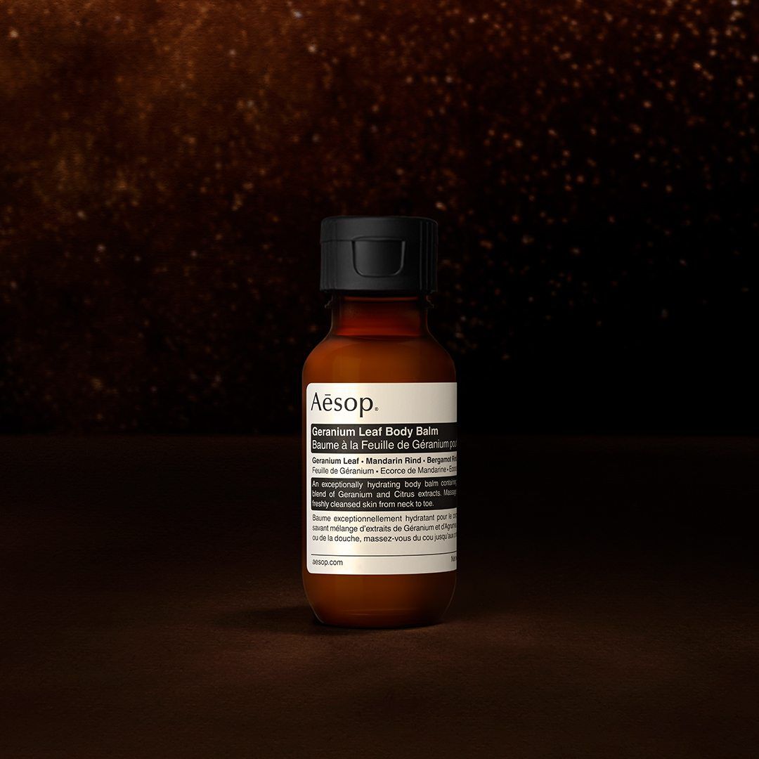 An Aesop Geranium Leaf body balm bottle against a dark, textured background.