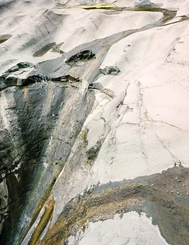 Pale stone and mineral surface with white, grey and charcoal striations, layered textures, and a diagonal fissure crossing the frame.